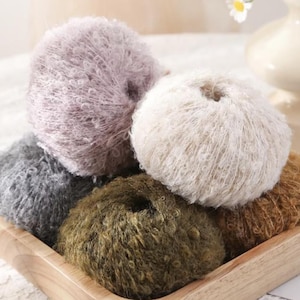 May include: A collection of fluffy yarn balls in various colors, including lavender, cream, gray, olive green, and brown. The yarn is arranged in a wooden tray, showcasing the soft texture and diverse color palette.
