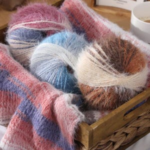 May include: A wooden tray holds several balls of fluffy yarn in shades of pink, blue, and brown. The yarn appears soft and textured, with a mix of colors. A knitted scarf with pink and purple stripes is also in the tray, suggesting a knitting or craft theme.