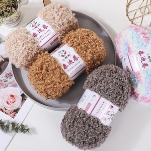 Peut inclure: Plusieurs pelotes de laine à tricoter "Teddy perm" de différentes couleurs, dont beige, marron et gris. Les pelotes sont disposées sur une assiette grise. Chaque pelote porte une étiquette avec des caractères chinois et "Teddy perm KNITTING".