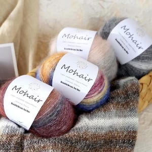 May include: A selection of Mohair yarn balls in a woven basket, alongside a knitted item. The yarn labels read "Mohair", and the balls display a range of colours, including pink, grey, and a blend of rainbow shades. The yarn is ideal for knitting.