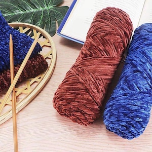K&ouml;nnte beinhalten: Nahaufnahme verschiedener Strickutensilien. Zwei gro&szlig;e Str&auml;nge aus weichem, strukturiertem Garn in Rost und Blau liegen neben einem kleinen Korb mit blauen und braunen Strickst&uuml;cken und zwei Stricknadeln aus Holz. Im Hintergrund ist ein Buch ge&ouml;ffnet.