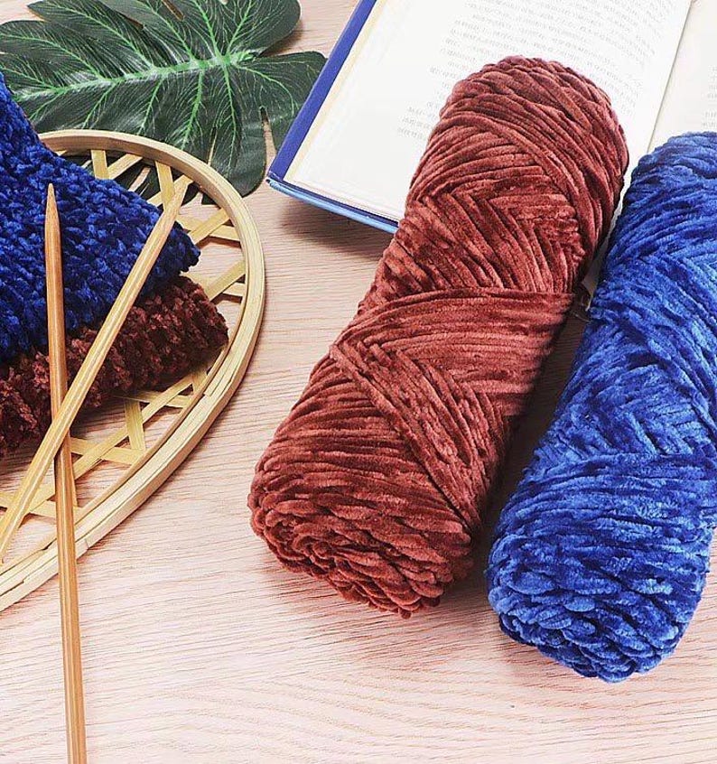 K&ouml;nnte beinhalten: Nahaufnahme verschiedener Strickutensilien. Zwei gro&szlig;e Str&auml;nge aus weichem, strukturiertem Garn in Rost und Blau liegen neben einem kleinen Korb mit blauen und braunen Strickst&uuml;cken und zwei Stricknadeln aus Holz. Im Hintergrund ist ein Buch ge&ouml;ffnet.