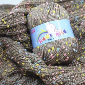 May include: A ball of brown yarn with colorful flecks of pink, yellow, and blue. The yarn is next to a knitted fabric of the same color and texture. The label on the yarn ball has text and a baby graphic.