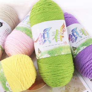 May include: A collection of colorful yarn skeins in shades of green, yellow, pink, purple, and beige. Each skein has a label with text and a butterfly graphic. The yarn is tightly wound, suggesting it's for knitting or crochet projects.