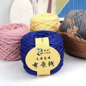 May include: A collection of colorful yarn balls in pink, yellow, blue, and gray. The blue yarn ball has a label with Chinese text. The yarn is thick and appears to be made of fabric strips, suitable for various crafting projects.