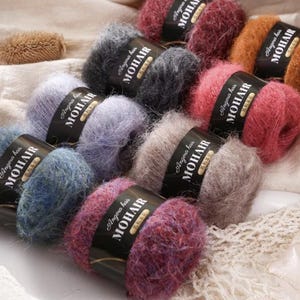 Op de afbeelding: Een verzameling kleurrijke mohair garenbollen in verschillende tinten, waaronder blauw, paars, roze en bruin. Elke bol is voorzien van het label "Angora hair MOHAIR" in witte tekst op een zwarte band. Het garen ziet er zacht en pluizig uit, ideaal voor brei- of haakprojecten.