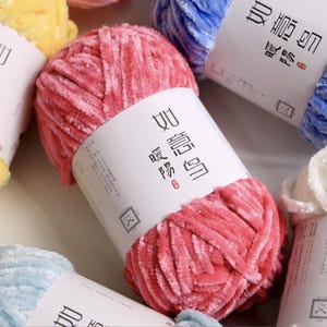 May include: Close-up of a ball of pink yarn with a white label featuring Chinese characters. The yarn has a soft, velvety texture. Other balls of yarn in various colors, including blue and yellow, are visible in the background.