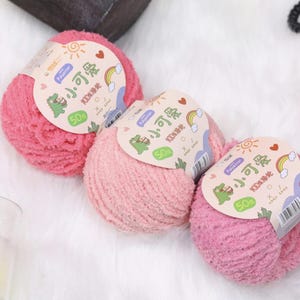 Könnte beinhalten: Drei Knäuel rosa Garn mit Etiketten mit einem Cartoon-Dinosaurier, Regenbogen und Sonne. Die Etiketten enthalten auch chinesische Schriftzeichen und den Text "Polyester" und "50%" auf weißem Hintergrund. Das Garn ist flauschig.
