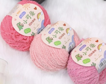 May include: Three balls of pink yarn with labels featuring a cartoon dinosaur, rainbow, and sun. The labels also include Chinese characters and the text "Polyester" and "50%" on a white background. The yarn is fluffy.
