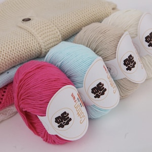 May include: A collection of colorful yarn skeins in pink, light blue, beige, and white, with a knitted sweater in the background. Each skein has a label with Chinese characters and the word "Cotton". The yarn is likely for knitting or crochet projects.