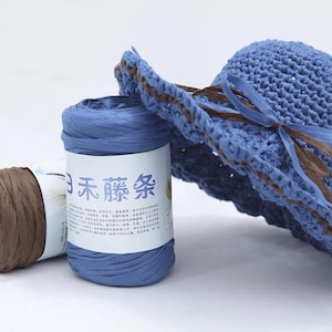 May include: A blue woven sun hat with a brown trim and bow, beside a roll of blue yarn and a roll of brown yarn. The blue yarn roll has text on the label.