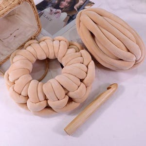 May include: A beige, braided yarn wreath and a large, oval-shaped skein of the same yarn. A wooden crochet hook is also present. The items are on a white surface, suggesting a craft or home decor theme.