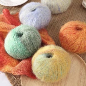 May include: A selection of colourful yarn balls in shades of green, orange, yellow, and blue. The yarn has a soft, fluffy texture, possibly mohair. The yarn is arranged on a wooden tray and a colourful scarf, ideal for knitting or crochet projects.