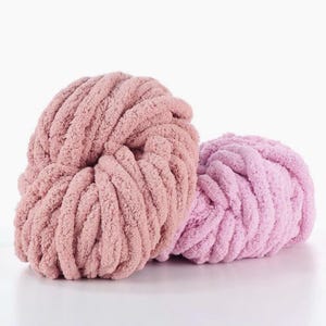 May include: Two balls of soft, chunky yarn in shades of pink and lavender. The yarn has a thick, looped texture, perfect for creating cozy blankets, scarves, or other handmade items. The yarn is displayed on a white background.