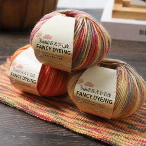 以下が含まれることがあります： 「FANCY DYEING」の英語と中国語の文字が書かれたラベルが付いた、カラフルな毛糸の3つの玉。毛糸は赤、黄色、グレーのミックスです。縞模様の生地の上に玉が置かれています。