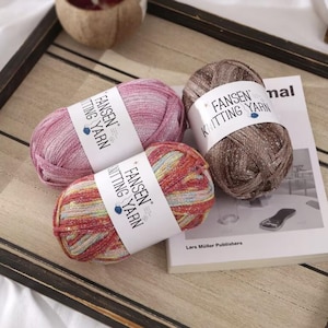 Könnte beinhalten: Drei Knäuel FANSEN Strickgarn in Rosa, Braun und Mehrfarbig. Jedes Knäuel hat ein weißes Etikett mit dem Markennamen. Das Garn wird auf einem Holztablett mit einem Buch im Hintergrund präsentiert.
