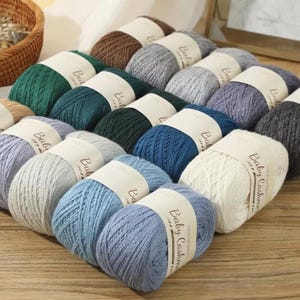 Op de afbeelding: Een verzameling garen in diverse kleuren, waaronder groen, blauw, grijs en bruin. Elk garen is verpakt met een papieren band met de tekst "Baby Cashmere". Het garen is netjes gerangschikt, wat duidt op een knutsel- of breibenodigdheid.
