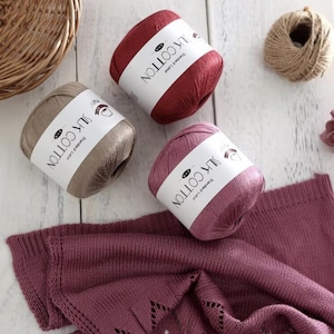 Könnte beinhalten: Drei Stränge Seidenbaumwollgarn in den Farbtönen Burgunder, Mauve und Taupe mit weißen Etiketten. Ein Strickstoff in passender Burgunderfarbe ist ebenfalls zu sehen. Eine Kordelrolle befindet sich im Hintergrund.