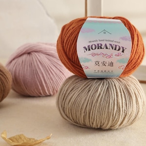May include: Three balls of yarn in shades of orange, pink, and beige are displayed. The orange ball has a label with "Moandy hand-knitted wool - MORANDY" in English and Chinese. The yarn is soft, ideal for knitting and other craft projects.