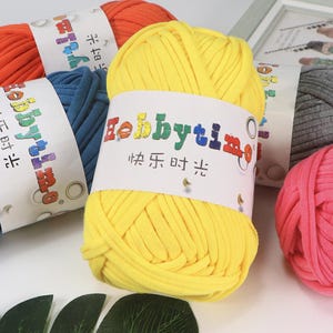Può includere: Primo piano di diversi gomitoli di lana colorati. Un gomitolo giallo brillante è al centro, con un'etichetta con la scritta "hobbytime". Altri gomitoli sono visibili in arancione, blu, grigio e rosa. La lana sembra essere fatta di un materiale morbido e spesso.