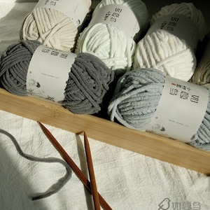 May include: A selection of yarn skeins in shades of grey, white, and pale green, presented in a wooden tray. Two wooden knitting needles are positioned near the yarn. The yarn skeins are labelled with text.
