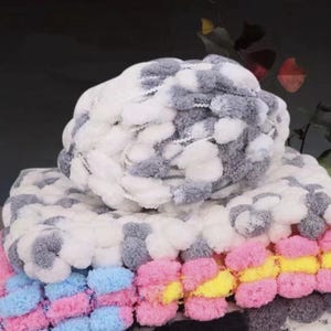 May include: A close-up of a soft, textured blanket. The blanket is made of white, grey, pink, yellow, and blue pom-poms. The pom-poms are arranged in rows, creating a cosy and colourful design. The blanket appears to be handmade.