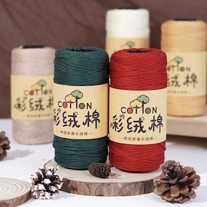 Könnte beinhalten: Spulen aus farbenfrohem Baumwollgarn in Grün, Rot, Beige, Weiß und Gelb. Jede Spule hat ein Etikett mit chinesischen Schriftzeichen und dem Wort "COTTON". Das Garn wird auf einer Holzoberfläche mit Tannenzapfen präsentiert.