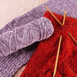 K&ouml;nnte beinhalten: Nahaufnahme von Strickzubeh&ouml;r. Ein Kn&auml;uel aus lavendelfarbenem Samtgarn liegt neben einem gestrickten Stoffmuster in leuchtendem Rot-Orange. Holzstricknadeln stecken im Strickstoff, was auf ein laufendes Projekt hindeutet.
