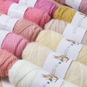May include: A selection of colourful yarn skeins in shades of pink, cream, and yellow. Each ball is labelled with a white band featuring a squirrel knitting illustration. The yarn appears soft and is suitable for knitting or crochet projects.