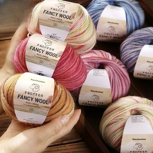 Puede incluir: Una colección de ovillos de lana Fancy Wool de colores. Las etiquetas presentan caracteres chinos y las palabras "FANCY WOOL". Los colores de la lana incluyen rosa, amarillo, naranja, azul y morado. La lana parece suave y esponjosa.