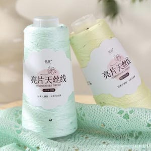 May include: Two spools of luminous silk thread in pastel shades. One is a light mint green, the other a pale yellow. Each spool has a white label with Chinese text and the words "Luminous Silk Thread". The thread is 100% silk.