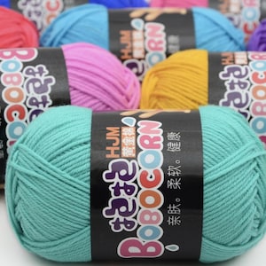 May include: A selection of colourful yarn skeins in shades of teal, pink, yellow, and blue. Each skein features a black label with white and pink text, including the word "BOBOCORN". The yarn appears soft, ideal for knitting or crochet.