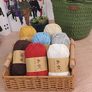 Op de afbeelding: Een rieten mand gevuld met kleurrijke garenbollen. Het garen is in tinten bruin, rood, geel, blauw, grijs en wit. Elke bol heeft een papieren label met Chinese tekst. Een groene gehaakte tas is op de achtergrond te zien.