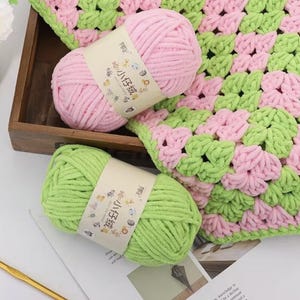 Op de afbeelding: Twee bollen roze en groen garen met labels, naast een gehaakte deken in bijpassende kleuren. Het garen is zacht en pluizig, perfect voor brei- of haakprojecten. De deken heeft een granny square ontwerp.