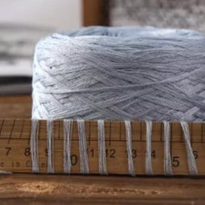May include: A large, light blue yarn ball sits behind a wooden ruler. The ruler has white threads wrapped around it, with measurements in inches. The yarn appears soft and is tightly wound. The background is blurred, suggesting a close-up shot.