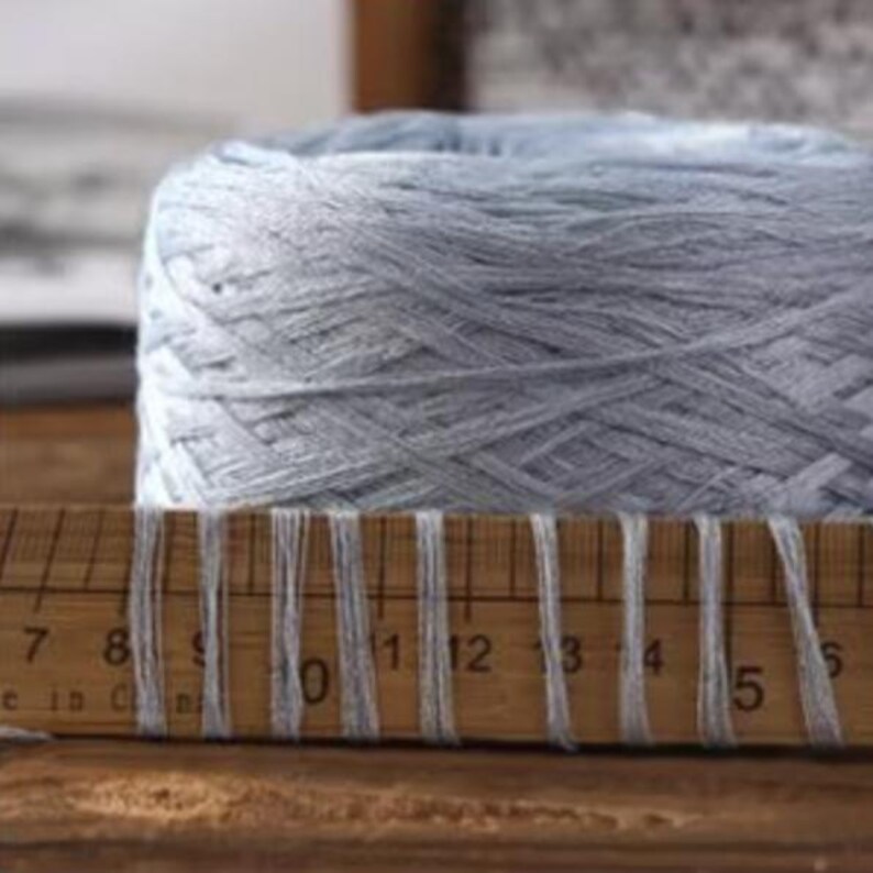 May include: A large, light blue yarn ball sits behind a wooden ruler. The ruler has white threads wrapped around it, with measurements in inches. The yarn appears soft and is tightly wound. The background is blurred, suggesting a close-up shot.