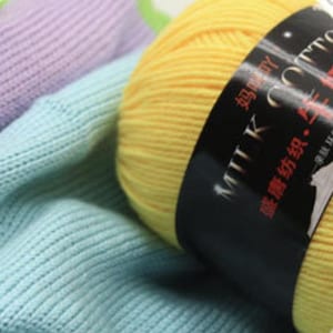 Può includere: Primo piano di gomitoli di filato colorati. Un gomitolo giallo brillante è a fuoco, con un'etichetta nera che recita "MILK COTTON" in bianco. Morbido tessuto a maglia blu e viola sullo sfondo, che suggerisce un tema artigianale.