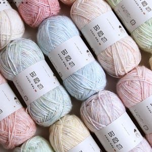 May include: A collection of yarn skeins in various pastel colors, including pink, blue, and yellow. Each skein is wrapped with a white label featuring text. The yarn appears soft and fluffy, suitable for knitting or crochet projects.