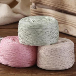 May include: Three skeins of yarn in pastel colors: light pink, pale green, and off-white. The yarn is tightly wound into round balls, suitable for knitting or crochet projects. The background includes a wooden crate and a white cloth.
