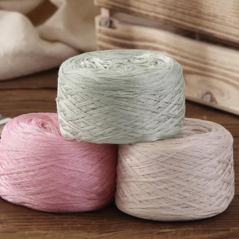 May include: Three skeins of yarn in pastel colors: light pink, pale green, and off-white. The yarn is tightly wound into round balls, suitable for knitting or crochet projects. The background includes a wooden crate and a white cloth.