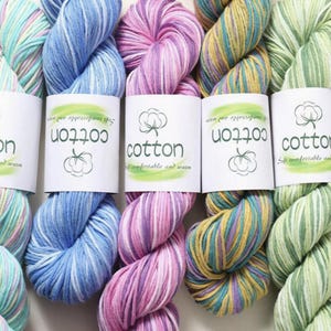 Op de afbeelding: Close-up van verschillende strengen kleurrijk katoenen garen. Het garen is in tinten blauw, roze, geel en groen. Elke streng heeft een wit label met het woord "cotton" en een katoenplantillustratie. De tekst "Soft comfortable and warm" staat ook op het label.
