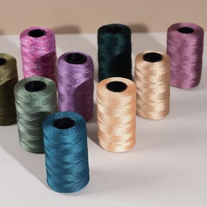 May include: A collection of colorful spools of thread in various shades, including pink, purple, green, and blue. The spools are cylindrical with a dark center, and the thread has a shiny, smooth texture. The arrangement suggests a craft or sewing theme.