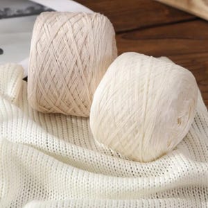 May include: Two skeins of yarn, one in a light beige and the other in white, rest on a cream-colored knitted fabric. The yarn is tightly wound, showing the texture of the fibers. The scene suggests a craft or textile project.