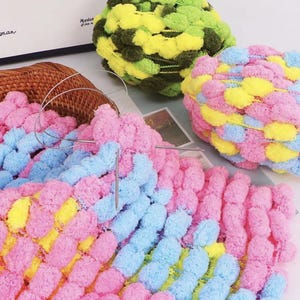 May include: A close-up shot of colourful yarn in pink, blue, and yellow, with a knitting needle inserted. The yarn is arranged in a pattern, with a basket and other yarn balls in the background. The yarn balls are green, yellow, and pink.