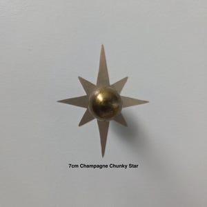 May include: A champagne-coloured star-shaped decorative object with a gold-coloured sphere at its centre. The star has eight points and is approximately 7 cm in diameter. The text "7cm Champagne Chunky Star" is below the star.