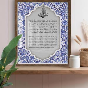 Puede incluir: Obra de arte enmarcada con caligrafía árabe en un borde floral decorativo azul y blanco. El texto está centrado sobre un fondo gris claro. El marco de madera añade un toque clásico a esta pieza religiosa o espiritual.