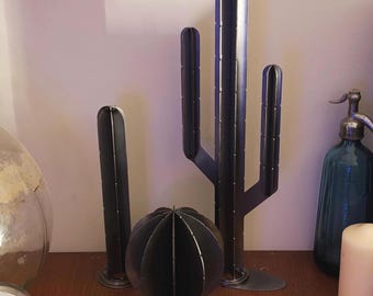 May include: Metal cactus sculptures in varying sizes and shapes. The largest is a tall, multi-stemmed cactus, while others are smaller, including a spherical design. All are dark gray and sit on a wooden surface, creating a modern desert theme.