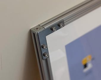 May include: Close-up of a framed artwork's corner. The frame is made of silver-colored metal with a rectangular profile. The corners are joined with metal brackets and secured with screws. The artwork has a white border and a blue background.