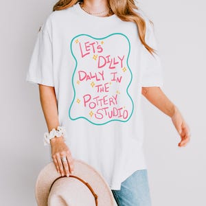 May include: A white short-sleeved t-shirt featuring a teal wavy-edged frame containing the pink text "LET'S DILLY DALLY IN THE POTTERY STUDIO" accented with small yellow star shapes. The design is centered on the front of the garment.
