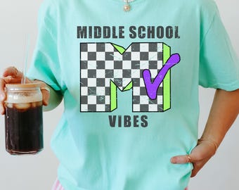 Może przedstawiać: Jasnozielona koszulka z krótkim rękawem z dużą grafiką na klatce piersiowej. Grafika przedstawia napis MIDDLE SCHOOL w kolorze ciemnoszarym, dużą literę M w czarno-białą szachownicę, fioletowy znaczek wyboru oraz napis VIBES.